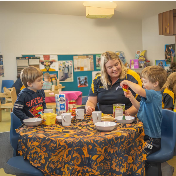 "This image portrays a warm and engaging environment in a childcare or early learning center. A woman, likely a caregiver or teacher, is seated at a table with two young children. They are involved in an interactive activity that involves food items. The children appear attentive and engaged, while the caregiver has a gentle smile on her face. The background is decorated with colorful artwork and educational materials, creating a vibrant learning space. 