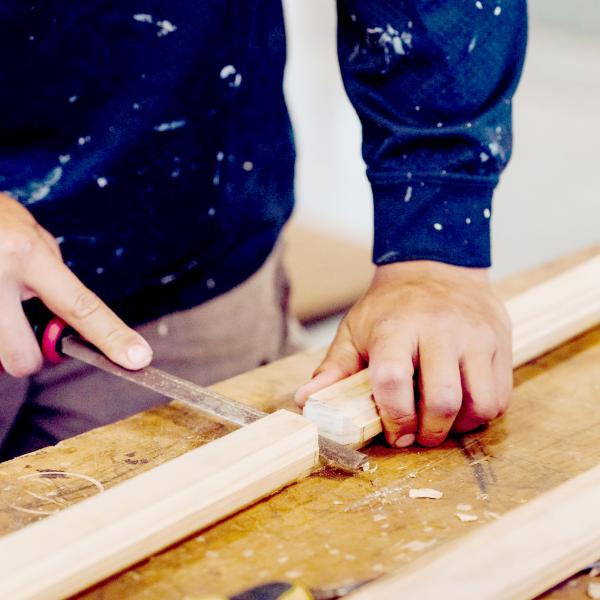 The image shows a close-up of a person working on a woodworking project. They are using a hand tool, likely a file or rasp, to shape or smooth a piece of wood. The individual's hands are firmly holding both the tool and the wooden piece, demonstrating precision and focus on the task.