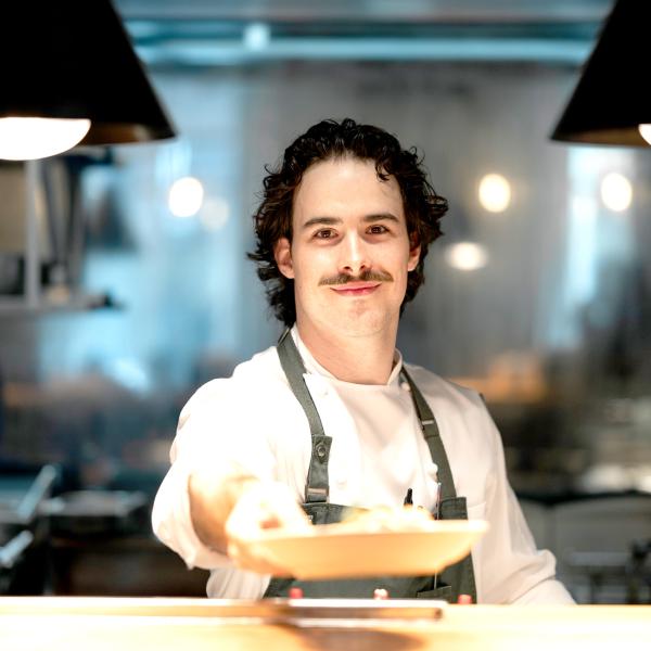 This image features another chef in a professional kitchen environment, presenting a dish with a friendly smile. The background includes kitchen equipment and a warm ambiance, highlighting the chef's dedication to culinary excellence and customer service.