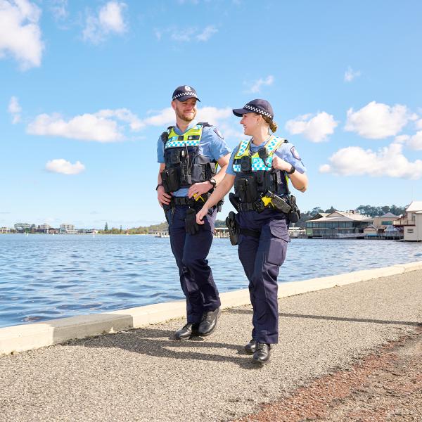The officers from the previous images are again seen walking, this time along a path closer to the water's edge. They seem engaged in a casual conversation, with the female officer smiling. The background includes a distant view of the city's skyline and a variety of modern and traditional architectural styles, adding to the city's coastal vibe.