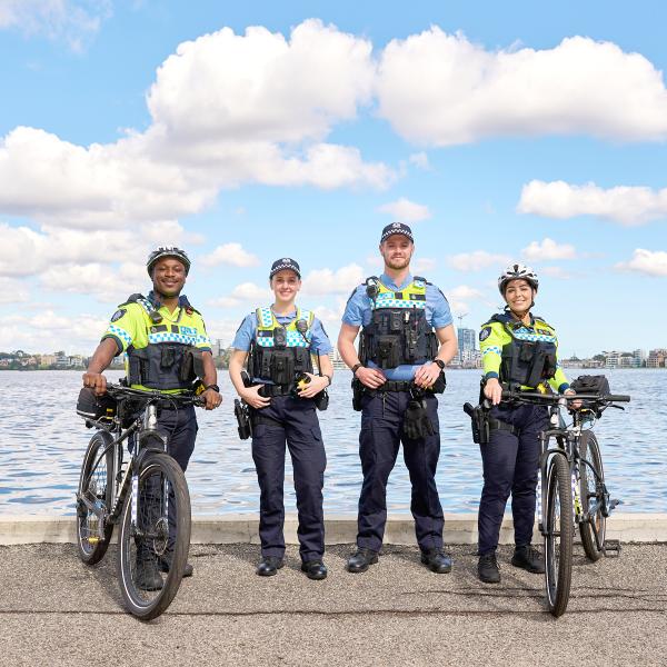 Four police officers are posing for a group photo near the waterfront. Two officers are on bicycles, while the other two are standing with their hands on their hips. They are wearing full police uniforms with tactical gear, including high-visibility vests and helmets. The sky is bright blue with scattered clouds, and the water is calm, providing a scenic and professional backdrop. The officers are smiling, indicating a sense of camaraderie and teamwork.